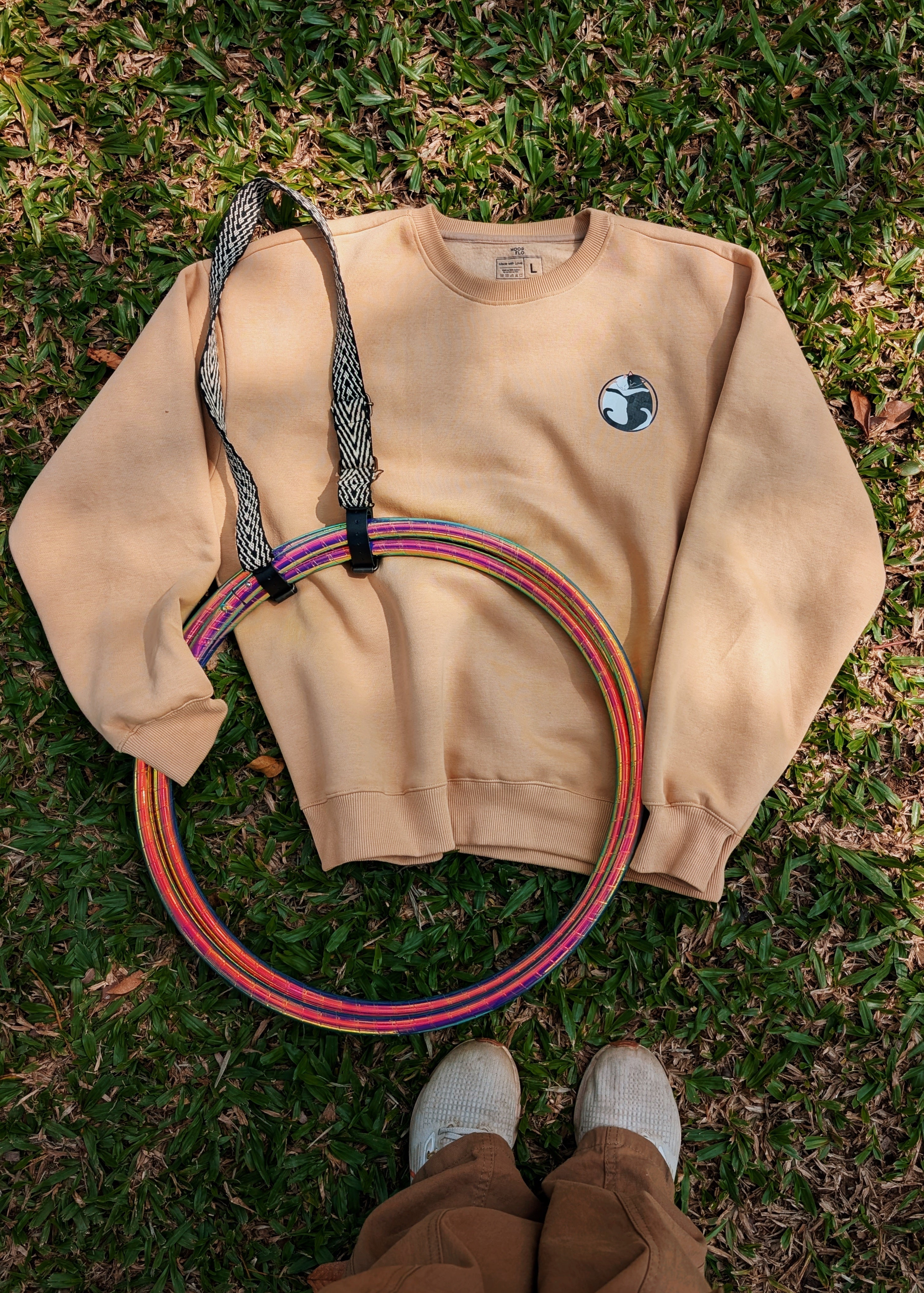 A chai-latte-coloured sweatshirt with a graphic of a yin-yang cat on the front, accompanied by a colorful hoop, placed on grass with a person's feet in pants visible in the foreground.